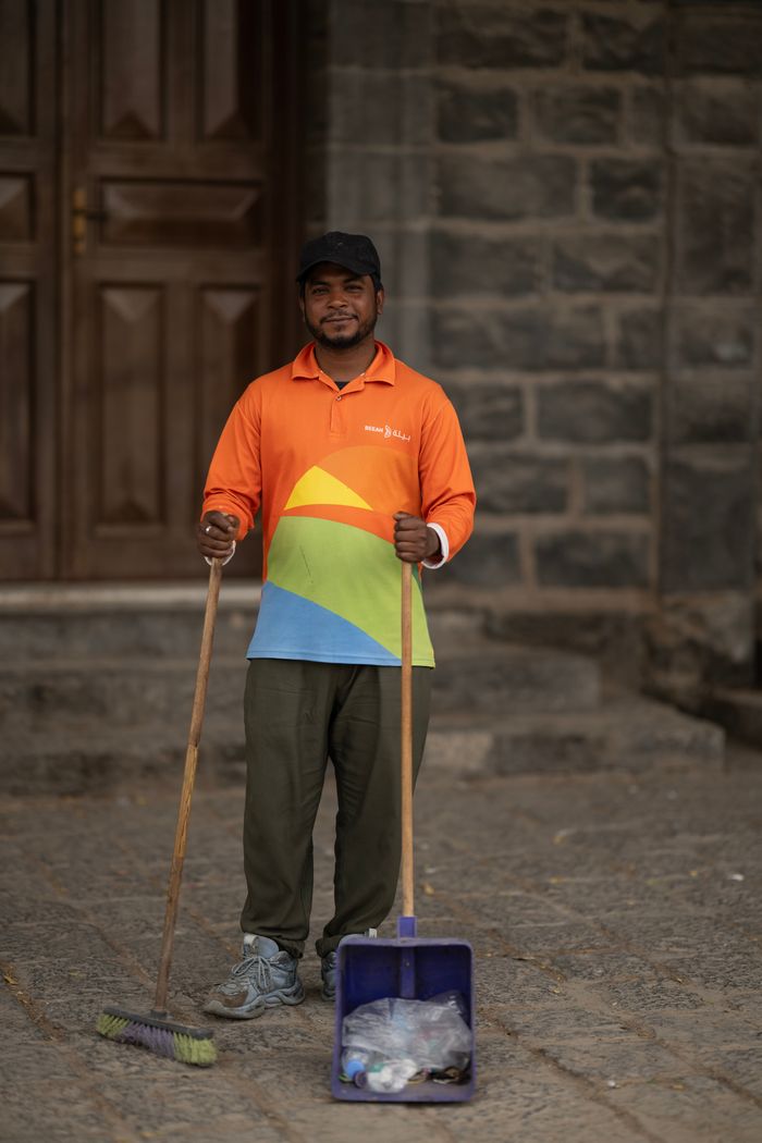 A street sweeper stands with broom and dustpan in front of stone building.